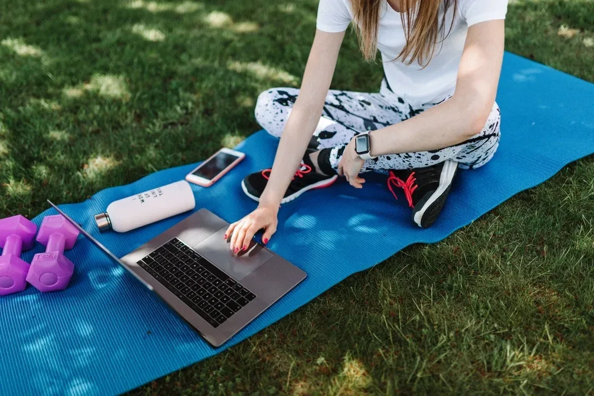 Woman in activewear using a laptop on a yoga mat outdoors, surrounded by fitness gear and technology.